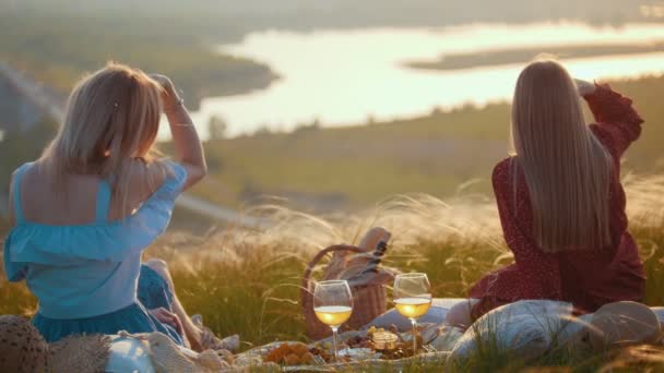 Zwei schöne Frauen sitzen auf dem Feld und picknicken - genießen einen Blick auf den Fluss bei Sonnenuntergang — Stockvideo