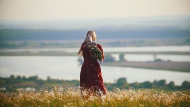 Een vrouw in rode jurk spelen met haar kleine baby op het veld op een achtergrond van de rivier — Stockvideo