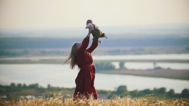 Een gelukkige vrouw spelen met haar kleine baby op het veld op een achtergrond van de rivier — Stockvideo