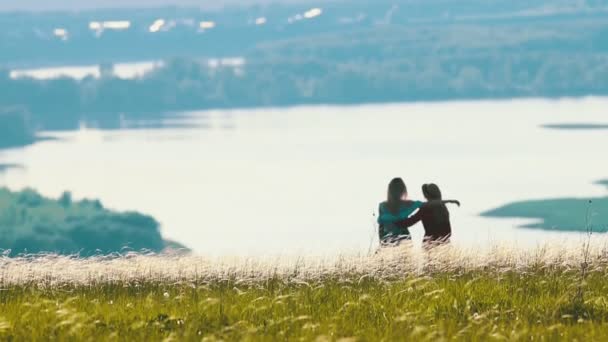 Dos mujeres jóvenes se abrazan en el campo en un día soleado. En el fondo del lago . — Vídeos de Stock