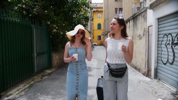 通りの間を歩いて笑っている二人の若い女性。そのうちの1人はスーツケースを運ぶ. — ストック動画