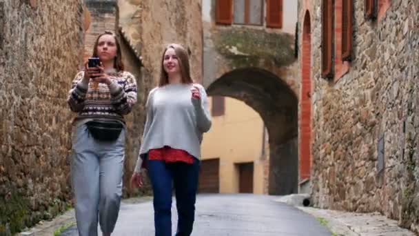 Две молодые женщины ходят по узкой улице и разговаривают. Один из них фотографирует. . — стоковое видео