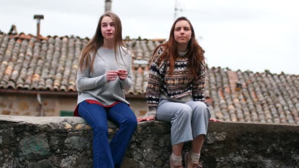 Dos mujeres jóvenes se sientan en una cornisa de hormigón y miran a su alrededor — Vídeos de Stock