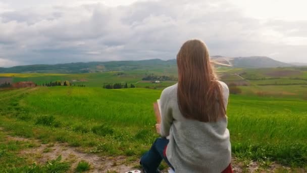 Mulher artista senta-se em uma colina em um campo verde e desenha — Vídeo de Stock