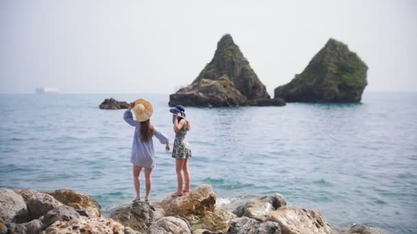 Две молодые женщины стоят на камнях в море и махают руками — стоковое видео