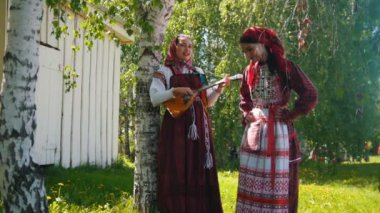 Rus elbiseli iki genç kadın çayırda duruyor - bunlardan biri balalaika oynuyor.