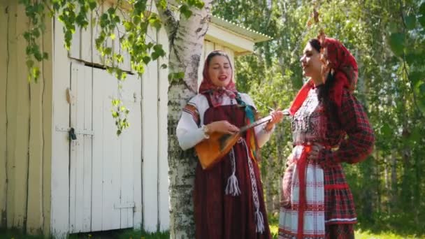 Dos mujeres jóvenes con ropa tradicional rusa de pie cerca de un edificio de madera una de ellas juega balalaika . — Vídeo de stock