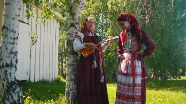 Two young women in russian dresses standing on the meadow - one of them plays the balalaika. — Stock Video