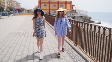 Elbiseler ve panama iki çekici kadın, sahil boyunca yürümek ve konuşurken
