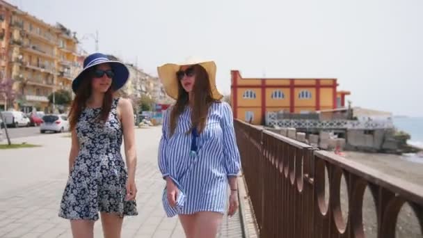 Две привлекательные женщины в платьях и панамах идут по улице рядом с пляжем — стоковое видео