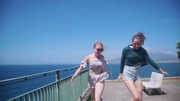 Zwei junge glückliche Frauen rennen und springen auf einer Aussichtsplattform vor dem Hintergrund des Meeres — Stockvideo
