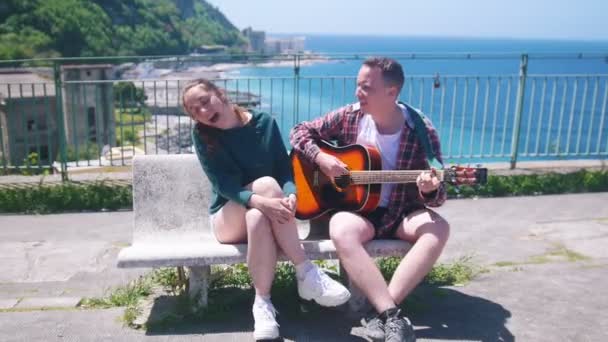 Um jovem casal sentado em um banco no deck de observação em um fundo da cidade resort perto do mar tocar guitarra e cantar — Vídeo de Stock