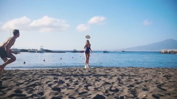 Молодой человек играет во фрисби на пляже со своей девушкой. Пытаясь поймать диск и упасть на песок — стоковое видео