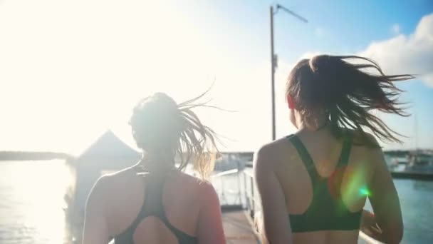Zwei junge Frauen mit Pferdeschwanz joggen bei sonnigem Wetter auf der Brücke — Stockvideo
