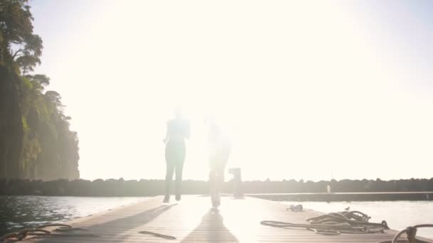 Dos mujeres jóvenes corriendo en el puente sobre el mar - luz solar brillante — Vídeo de stock