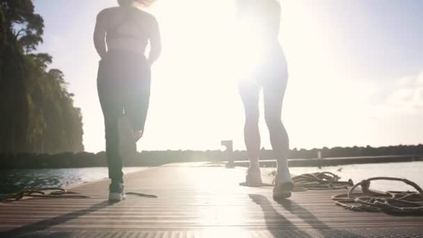 Dos mujeres jóvenes comienzan a trotar en el puente sobre el mar la luz del sol brillante — Vídeo de stock