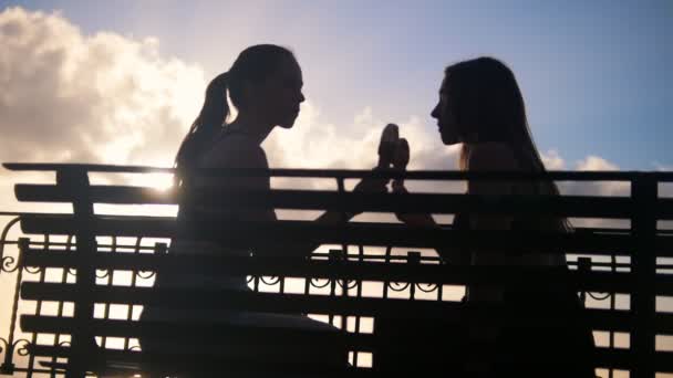 Два силуэта молодых женщин, сидящих на набережной и поедающих мороженое - дегустация друг друга мороженое — стоковое видео