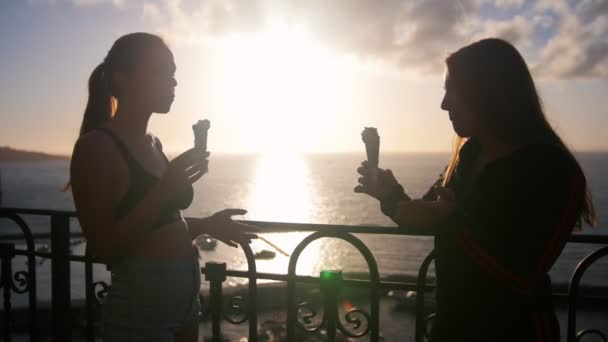 Zwei Silhouetten junger sprechender Frauen, die am Kai stehen und bei Sonnenuntergang Eis essen — Stockvideo