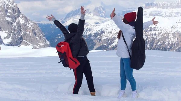 Duas jovens mulheres animadas de pé na neve em Dolomites com grande mochila e guitarra — Fotografia de Stock