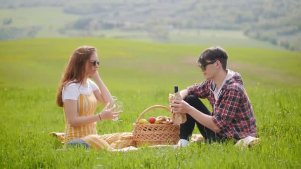Ett ungt par i ljusa kläder sitter på en ljus grön äng och hälla vin i glasen — Stockvideo