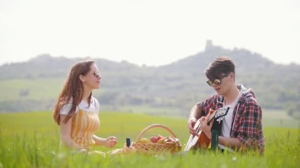 Ett ungt par i ljusa kläder sitter på en ljus grön äng och med en picknick-en man som spelar gitarr — Stockvideo