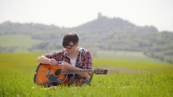 Ein junger Mann sitzt mit einer Gitarre auf einer hellgrünen Wiese und zündet sich eine Zigarette an — Stockvideo