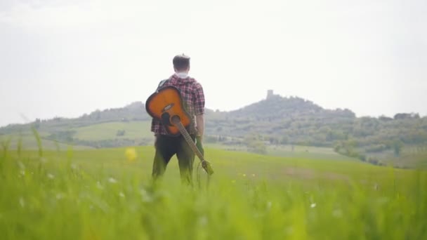 Parlak yeşil bir çayırda yürüyen genç bir gitarist - seyahat konsepti — Stok video