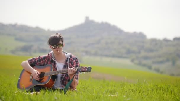 Egy fiatal férfi ül egy fényes zöld réten gitárral és cigarettázás — Stock videók