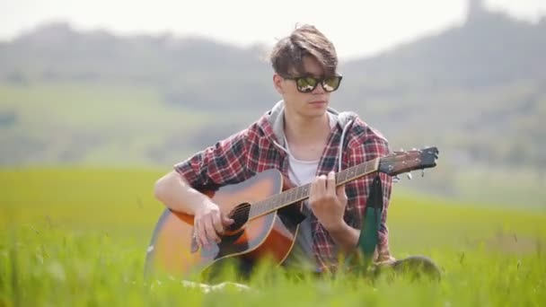 Молодой человек сидит на ярко-зеленом лугу и играет на гитаре — стоковое видео