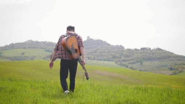 Parlak yeşil bir çayırda yürüyen genç bir gitarist — Stok video