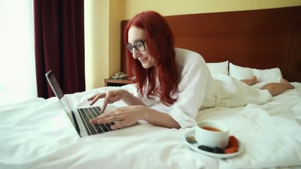 Une rousse couchée sur le lit et travaillant avec un ordinateur portable - travail indépendant — Video