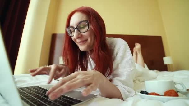 Una mujer pelirroja sonriente acostada en la cama y trabajando con un ordenador portátil - trabajo independiente — Vídeo de stock