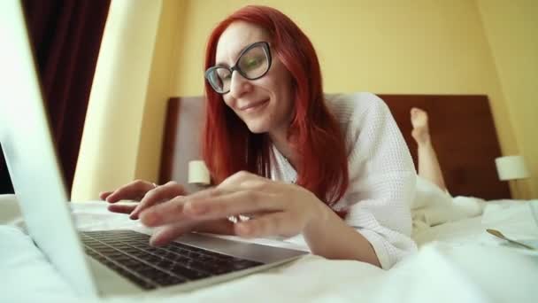 Una mujer pelirroja con gafas acostada en la cama y trabajando con un ordenador portátil - trabajo independiente — Vídeo de stock
