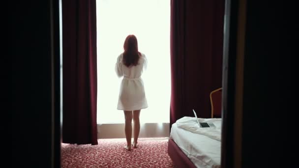 A ginger woman in bathrobe opens the curtains in hotel room and looking in the window — Stock Video