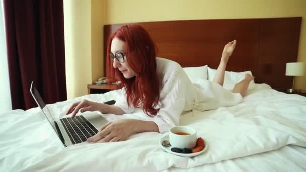 Імбирна жінка лежить на ліжку і працює з ноутбуком - позаштатна робота — стокове відео