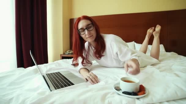 A gyömbéres nő feküdt az ágyon, és dolgozik egy laptop és tea-külsős munka — Stock videók
