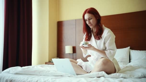 Uma mulher de gengibre sentada na cama no quarto do hotel - bebendo chá - trabalhando com um laptop — Vídeo de Stock