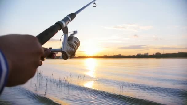 Un hombre en la pesca - girar la bobina en la caña de pescar - puesta de sol — Vídeo de stock