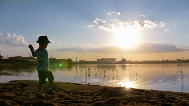 Un niño pequeño caminando hacia el agua y arrojando una roca al agua — Vídeos de Stock