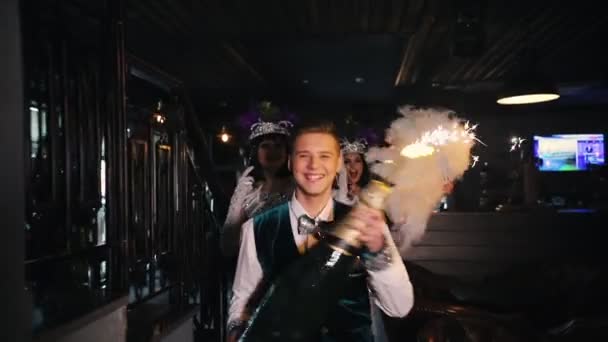 Retro party tematyczne-młody człowiek chodzenie towarzyszy dwie kobiety w genialny ubrania-trzymając ogromną butelkę szampana z fajerwerkami na szczycie — Wideo stockowe
