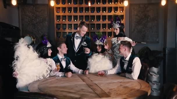 Theme party - People in glistening clothes sitting behind the table and talking - A man in the centre raises a glass of drink up — Stock Video