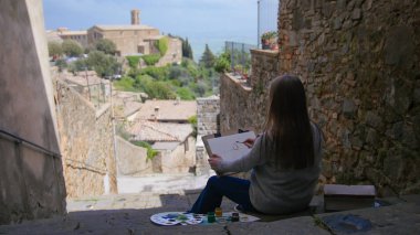 Genç kadın sanatçı merdivenlerde oturup eski binaları çiziyor