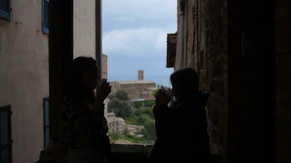 Силуэты двух молодых женщин стоят на балконе между зданиями и пьют кофе — стоковое фото
