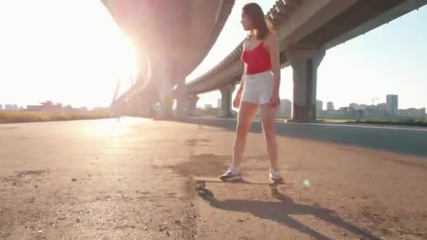 Mujer joven en pantalones cortos blancos montando un monopatín debajo del puente - camina fuera del tablero — Vídeo de stock