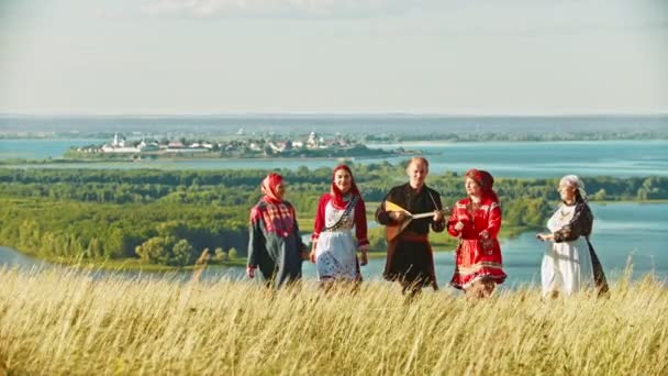 Människor i traditionella ryska kläder går på fältet och sjunger en sång-en man som innehar Balalaika — Stockvideo