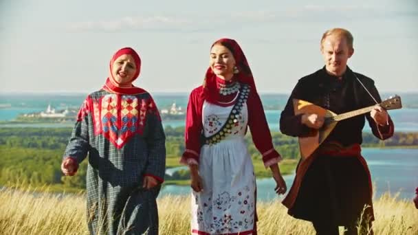 Lidé v tradičních ruských šatech, kteří chodí po hřišti a zpívají píseň-muž hrající balalajka — Stock video