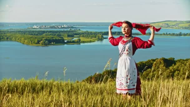 Eine junge Frau in russischer Tracht steht auf dem Feld und blickt auf den Fluss - Taschentuch flattert im Wind — Stockvideo