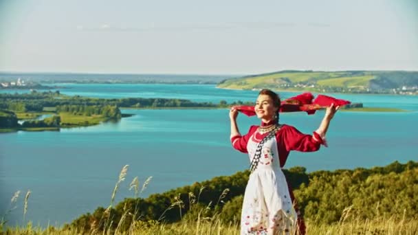 Een jonge lachende vrouw in Russische folk kleren staan op het veld op een achtergrond van de rivier-zakdoek wappert door de wind — Stockvideo