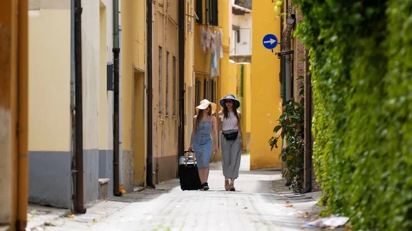 Deux jeunes femmes en lunettes de soleil et panamas marchant le long de la rue étroite et parlant — Photo