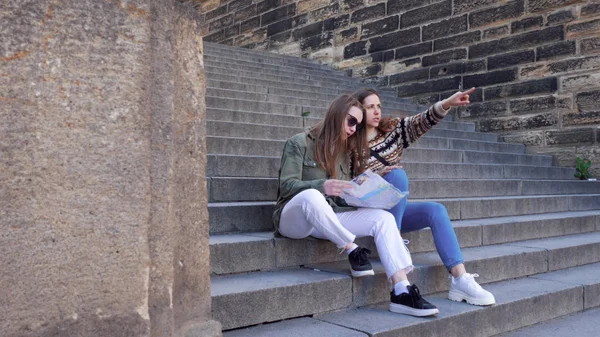 Dos mujeres jóvenes se sientan en las escaleras de ladrillo y miran en el mapa —  Fotos de Stock